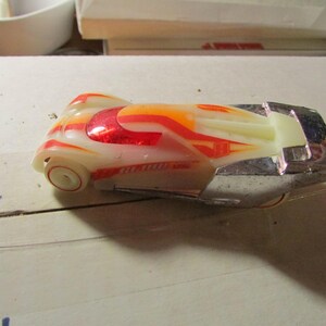 May include: A vintage toy car with a cream-colored body and vibrant red, orange, and yellow flame-like designs. The car has a chrome-colored base and wheels, and is set against a neutral background.