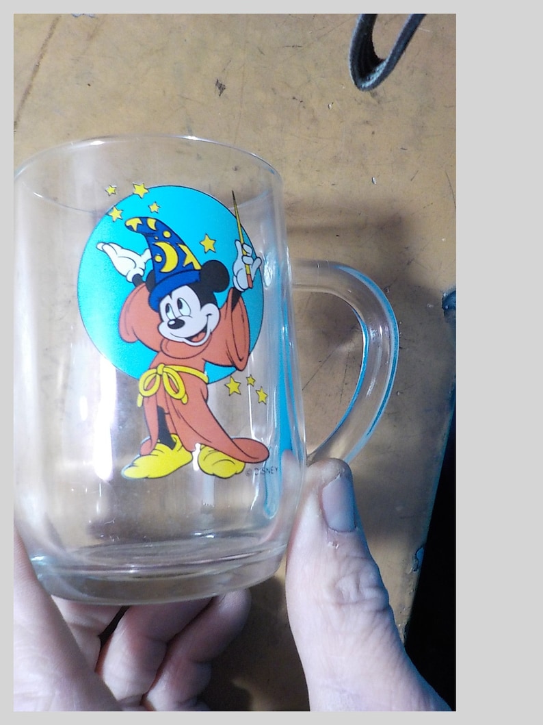 May include: Clear glass mug featuring a colorful graphic of Mickey Mouse as a sorcerer, with a blue hat and red robe. The mug has a handle and is likely for beverages.