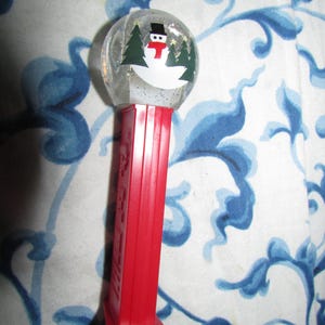 May include: A red and clear PEZ dispenser featuring a snowman design inside a snow globe. The snowman has a black top hat, a red scarf, and is surrounded by green trees. The dispenser is a festive holiday item.