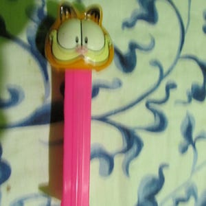 May include: A Garfield PEZ dispenser featuring the cartoon cat's face in orange, white, and black. The dispenser has a bright pink stem and is set against a patterned background.