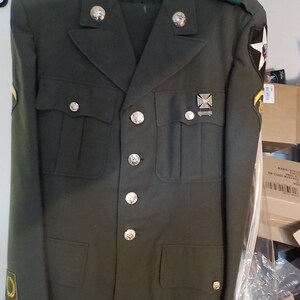 May include: Olive green military uniform jacket and pants set. The jacket features silver buttons, shoulder epaulets, and various patches and insignia. The uniform is hanging on a wooden hanger.