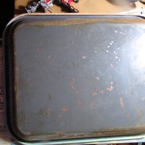 Puede incluir: Una bandeja rectangular de metal gris con esquinas redondeadas y un borde ligeramente elevado. La bandeja muestra signos de edad con manchas de color &oacute;xido y desgaste en los bordes. La bandeja es probablemente vintage.