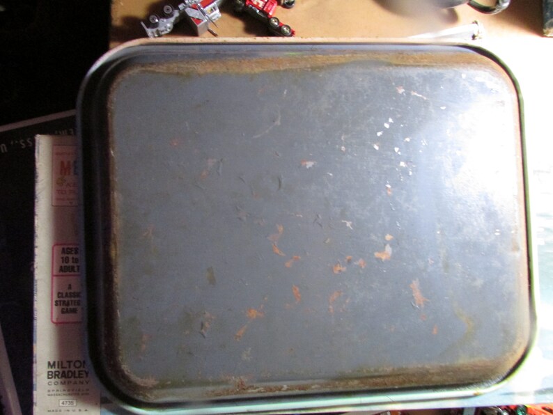 Puede incluir: Una bandeja rectangular de metal gris con esquinas redondeadas y un borde ligeramente elevado. La bandeja muestra signos de edad con manchas de color &oacute;xido y desgaste en los bordes. La bandeja es probablemente vintage.