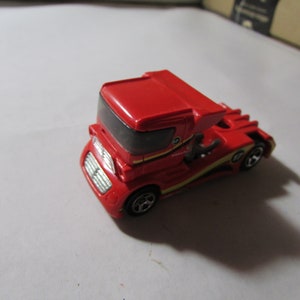May include: A red toy semi-truck with a black windshield and yellow accents. The toy truck has a detailed front grill and a unique aerodynamic design. The wheels are black and silver. This toy truck is a collectible item.