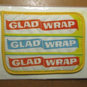 May include: A rectangular quilted fabric potholder with rounded corners and yellow trim. The white surface features three horizontal bands with the text GLAD WRAP printed in white on red and blue backgrounds, repeating the brand name three times.