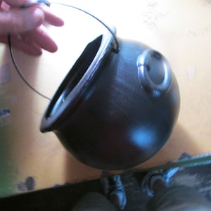 May include: A black, round cauldron with a handle and a small loop on the side. The cauldron has a wide opening and a curved shape. The handle is made of wire. The cauldron is likely used for cooking or decoration.