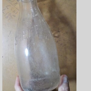 May include: A clear glass milk bottle with the embossed text "SHEFFIELD" on the front. The bottle has a rounded shape and a narrow neck. The bottle is held in a hand.
