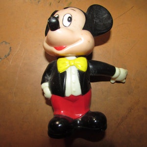 May include: A vintage Mickey Mouse figurine. The figure is wearing a black tuxedo jacket, red pants, a yellow bow tie, and black shoes. The figure has a white face, black ears, and a red smile.