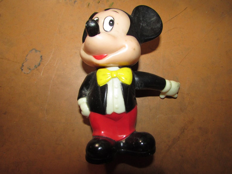 May include: A vintage Mickey Mouse figurine. The figure is wearing a black tuxedo jacket, red pants, a yellow bow tie, and black shoes. The figure has a white face, black ears, and a red smile.