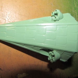 May include: A light green, angular toy spaceship with a textured surface. The toy has multiple circular protrusions on the top and sides, suggesting weaponry or engines. The design is reminiscent of a science fiction vehicle.