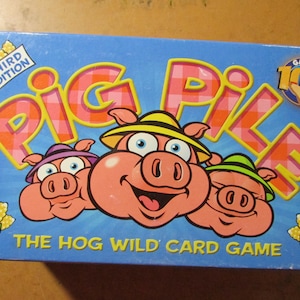 May include: A blue game box for "Pig Pile" card game. The box features cartoon pigs wearing hats, the game title in large letters, and the text "The Hog Wild Card Game". The box also has a "Third Edition" logo.