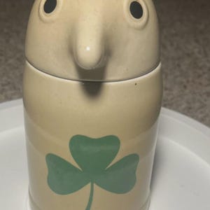 May include: A beige ceramic jar with a lid featuring a protruding nose, two black circular eyes, and brown arched eyebrows. The cylindrical body of the jar is decorated with a large, centered green shamrock graphic on the front surface.