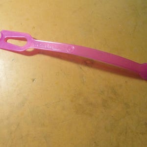 May include: A curved, pink plastic tool with a handle and a notched end. The tool has a hole near the notched end and the text "The Fine Touch" is printed on the handle. The tool is lying on a yellow surface.