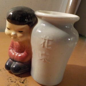 May include: A ceramic figurine of a kneeling child in red and black clothing, next to a light blue vase. The vase has embossed Chinese characters. The figurine has black hair and a serene expression. The vase is slightly curved.