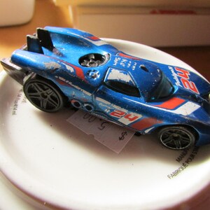 May include: A blue and red toy race car with the number 24 on the side. The car has black wheels and a weathered appearance. The toy car is sitting on a white surface.