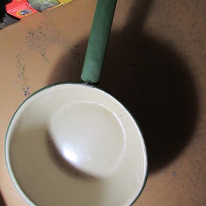 May include: A vintage, cream-colored saucepan with a green handle and rim. The handle has a red hole at the end. The pan is empty and appears to be made of enamel. The pan is sitting on a brown surface.