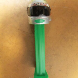 May include: A green and silver PEZ dispenser featuring a racing helmet design. The helmet has a black visor and the word "INTERSTATE" printed on the top. The dispenser is made of plastic.