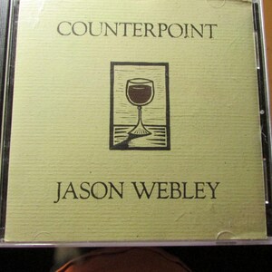 May include: A CD case featuring a light tan cover with the text COUNTERPOINT at the top and JASON WEBLEY at the bottom. In the center is a dark brown graphic illustration of a wine glass inside a rectangular frame on a flat surface.