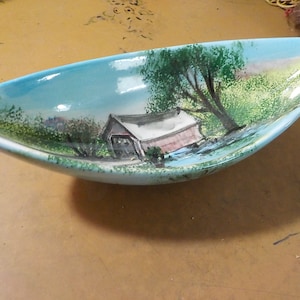 May include: A vintage, oblong ceramic dish with a hand-painted landscape scene. The interior features a covered bridge, trees, and a river against a light blue sky. The dish has a light blue rim and a glossy finish.