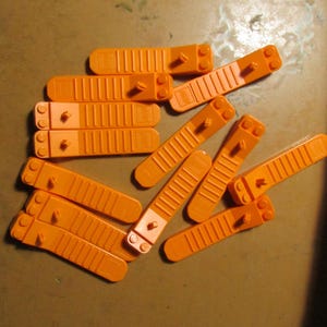 May include: A collection of orange plastic building block pieces. The pieces are rectangular with a series of parallel lines and a small peg on one end. The blocks are scattered on a brown surface.