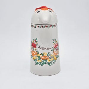 May include: A white ceramic lidded jar featuring a sculpted bird-like head on the lid with a red crest. The body is decorated with a hand-painted floral wreath surrounding the word Andenken in script, with green, orange, and red floral accents.