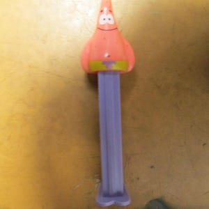 May include: A Patrick Star PEZ dispenser. The dispenser features a pink Patrick Star head with a smiling face, a yellow and green belt, and a purple stem. The dispenser is made of plastic.