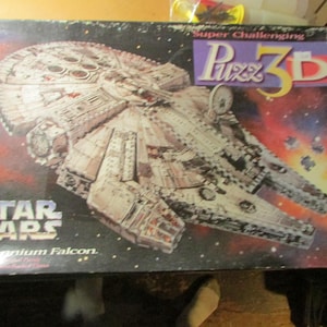May include: A rectangular box for a Star Wars Millennium Falcon Puzz 3D puzzle. The box features a large illustration of the spaceship against a dark space background with stars and nebula colors. The text reads Super Challenging Puzz 3D.