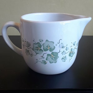 May include: White ceramic creamer with a handle and a spout. The creamer is decorated with a green and gold ivy leaf pattern. The creamer is sitting on a black surface.