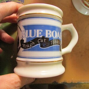 May include: A white ceramic mug with a blue and gold design. The mug features the words "BLUE BOARD Rough Cut Tobacco" in a stylized font. The mug has a handle and a gold trim around the top and bottom.