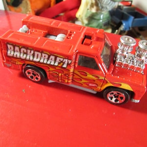 May include: A red toy truck with the word "BACKDRAFT" in white lettering on the side. The truck has flames painted on the side and a chrome engine. The toy truck is on a red surface.