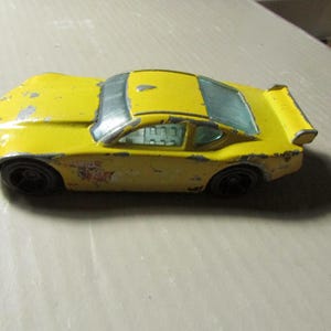May include: A vintage yellow toy race car with black wheels and a rear spoiler. The car has a weathered appearance with visible wear and tear. The windows are a light blue color. The car has a sticker on the side.