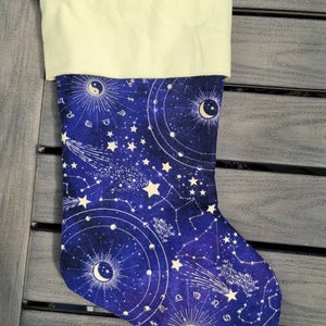 May include: A Christmas stocking with a celestial design. The stocking features a deep blue background with white constellations, stars, and zodiac symbols. The cuff is a light cream color, and a purple ribbon is attached for hanging.