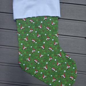 May include: A Christmas stocking featuring a green fabric body with a repeating pattern of bees wearing red Santa hats, interspersed with small red berries and green holly leaves. The stocking has a wide, solid white cuff and a red ribbon loop.
