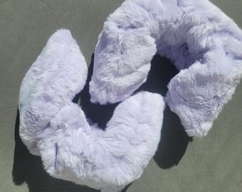 May include: A lavender-colored, plush neck pillow and eye mask set. The set is made of soft, faux fur material. The neck pillow is U-shaped, and the eye mask is a curved shape. The items are designed for comfort and relaxation.