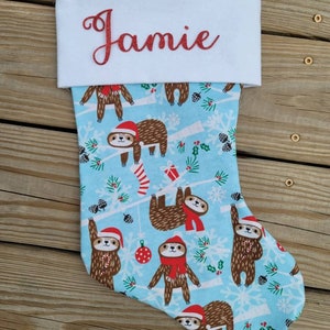 May include: A blue and white Christmas stocking with a sloth pattern. The stocking has a white cuff with the name "Jamie" embroidered in red glitter.