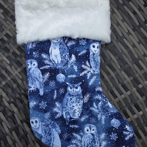 May include: A Christmas stocking with a blue background featuring white owls, snowflakes, and pine branches. The top of the stocking has a white faux fur trim and a white loop for hanging.