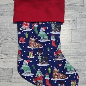 May include: A Christmas stocking featuring a dark blue fabric base with a repeating pattern of cats wearing holiday sweaters, scarves, and hats, interspersed with gift boxes and mistletoe. The stocking has a solid red cuff and a red hanging loop.