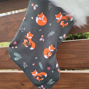 May include: A dark green Christmas stocking with a white faux fur cuff. The stocking features a repeating pattern of orange foxes with their kits, surrounded by small green leaves and red berries.