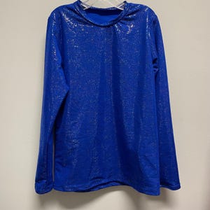 May include: A long-sleeved, royal blue shirt with a textured, shimmering surface. The shirt has a crew neck and is hanging on a white hanger. The fabric has a subtle, metallic sheen, adding a touch of visual interest to the garment.
