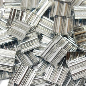 May include: A collection of small, square, silver mosaic tiles. Each tile has a reflective, striped surface, creating a shimmering effect. The tiles are scattered, showing their individual details and textures. Ideal for craft projects.
