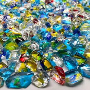 May include: A collection of colorful, irregularly shaped glass pieces. The glass is primarily shades of blue, green, yellow, and red, with clear and reflective surfaces. The pieces vary in size and are scattered across the image, creating a textured appearance.