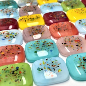 Op de afbeelding: Een verzameling kleurrijke vierkante glazen tegels met een gespikkeld patroon. De tegels zijn in rijen gerangschikt en hebben verschillende kleuren, waaronder blauw, groen, roze, rood en geel.