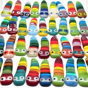 以下が含まれることがあります： カラフルで気まぐれなガラスアート作品のコレクション。各作品は、丸みを帯びた頭と平らなベースを持つユニークな細長い形状をしています。デザインはさまざまな色の水平ストライプを特徴とし、それぞれに目と口のある顔があります。