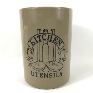 Puede incluir: Un porta utensilios de cerámica marrón con letras negras que dicen "Kitchen Utensils". El porta utensilios presenta un gráfico de tres utensilios de cocina.