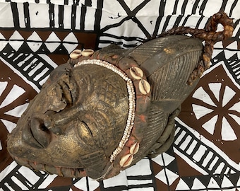 May include: A wooden mask with a carved face, closed eyes, and a beaded headdress with cowrie shells. The mask is painted in shades of brown and gold and has a textured surface.