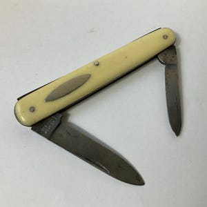 Puede incluir: Un pequeño cuchillo de bolsillo de dos hojas con un mango de plástico blanco. El cuchillo tiene una hoja simple y una hoja secundaria más pequeña. El mango tiene una incrustación ovalada plateada.