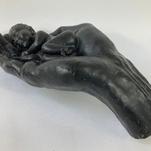 May include: A black sculpture of a hand cradling a sleeping baby. The sculpture is made of a smooth, matte material and has a realistic appearance.