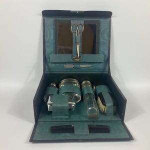 May include: An open, vintage black leather travel grooming kit with a teal interior. The kit contains a mirror, scissors, a nail file, bottles, a brush, and a comb. The kit is designed to hold grooming essentials.