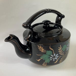 May include: A black ceramic teapot with a curved spout and a rounded body. The teapot features a black handle and lid, and is decorated with floral designs in green, blue, and gold. The teapot is designed for brewing tea.