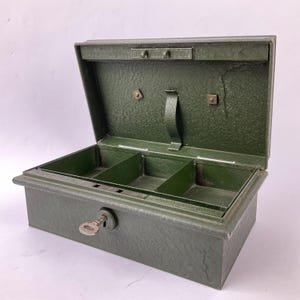 Op de afbeelding: Een rechthoekige olijfgroene metalen geldkist met een getextureerde afwerking en een open deksel. Het interieur is voorzien van een uitneembaar bakje met drie vakken. Een kleine metalen sleutel zit in het slot aan de voorzijde.
